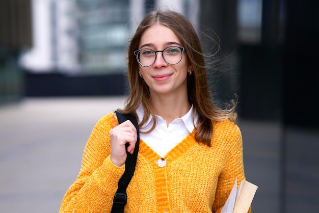 Portrait of happy cute girl, young blonde European woman university or college student in glasses with backpack and book is smiling, looking at camera outdoors at campus. Education, study concept.