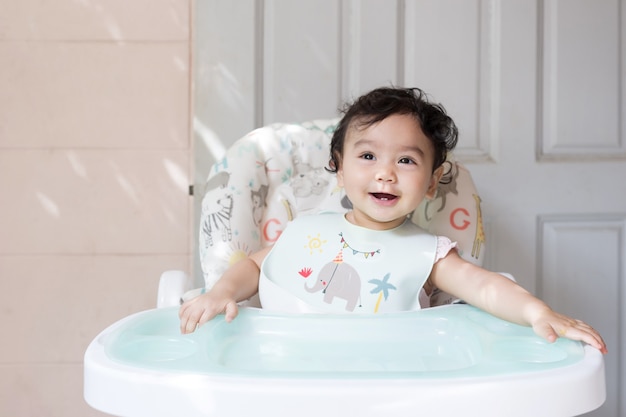 Portrait of happy cute little Asian baby girl siting on baby dining table smiling and looking at camera, baby expression concept