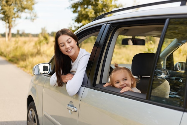 Portrait of happy delighted female with dark hair sitting in car with her baby daughter on back seat family looking through automobile window expressing positive emotions