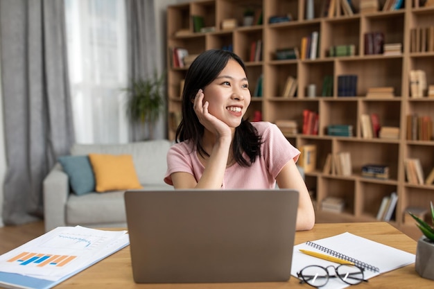 Portrait of happy dreamy asian lady sitting at desk with laptop at home looking away thinking and