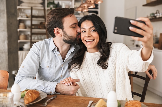 Portrait of happy european couple man and woman taking selfie photo while having breakfast in kitchen at home