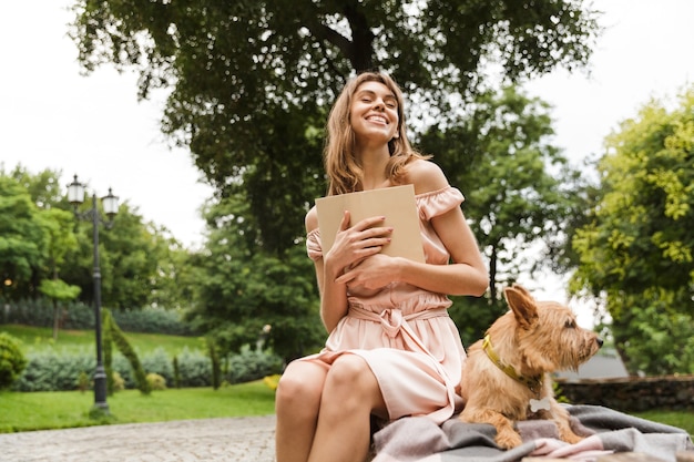 Portrait of happy excited woman wearing dress reading book while sitting on bench with her dog in a park