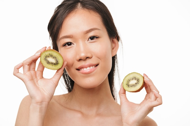 Portrait of a happy excited young beautiful asian woman posing isolated over white wall holding kiwi