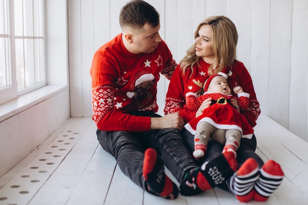 Portrait of happy family mom dad and little daughter in red traditional christmas clothes spending time together in light wooden room near window copy space