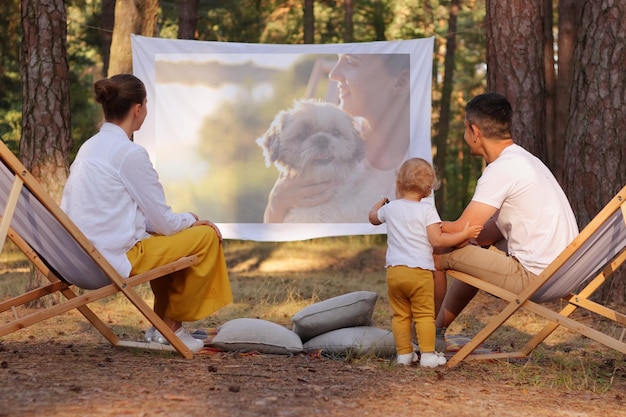 Portrait of happy family sitting in the forest on deck chairs with their little daughter and watching movie on projector parents playing with their cute kid and enjoying nature and fresh air