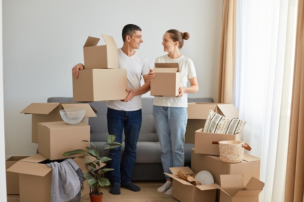 Portrait of happy family woman and man wearing white t shirts standing with cardboard boxes relocating to a new house unpacking personal belongings looking at each other with love real estate