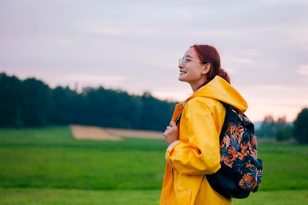 Portrait of a happy female tourist with a backpack in glasses wearing yellow coat. Green field