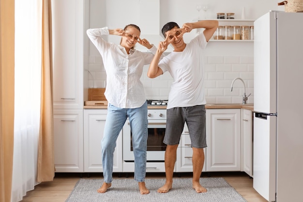 Portrait of happy funny family dancing in kitchen at home, making v sign gesture near eyes, smiling, celebrating completion of the repair in the house.