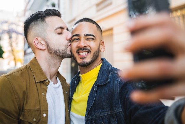 Portrait of happy gay couple spending time together and taking a selfie with mobile phone in the street. Lgbt and love concept.