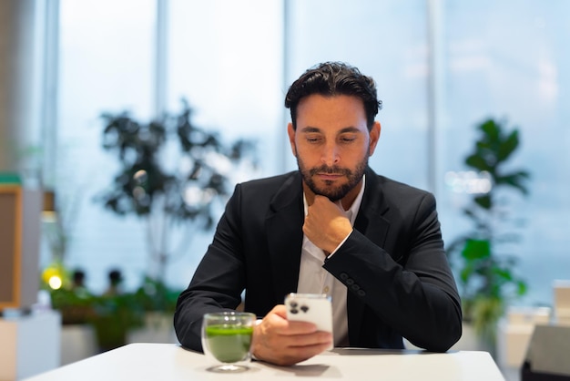 Portrait of happy handsome Hispanic businessman at coffee shop having green tea and using phone