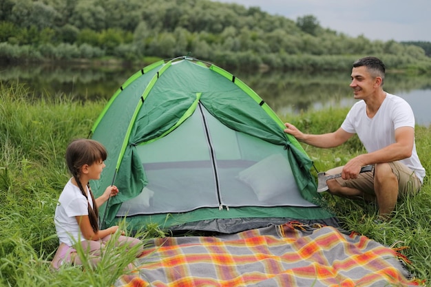 Portrait of happy handsome man wearing casual clothing posing near green tent with his cute daughter family having camping together enjoying beautiful nature