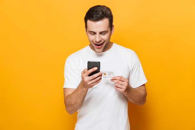 Portrait of a happy handsome man wearing t-shirt standing isolated over yellow wall, using mobile phone, showing credit card