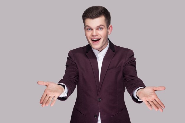 Photo portrait of happy handsome young man in violet suit and white shirt, standing, looking at camera with raised arms and toothy smile. indoor studio shot, isolated on grey background.