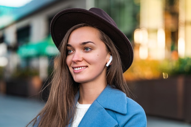 Portrait of happy joyful stylish trendy fashionable hipster girl in hat with wireless white headphones enjoys music in the city centre. Modern peoples lifestyle and technology