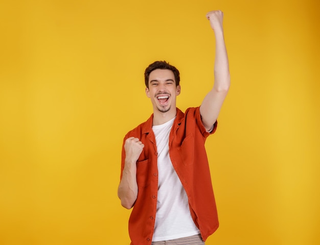 Portrait of happy joyful young man standing doing winner gesture clenching fists keeping isolated on yellow color wall background studio