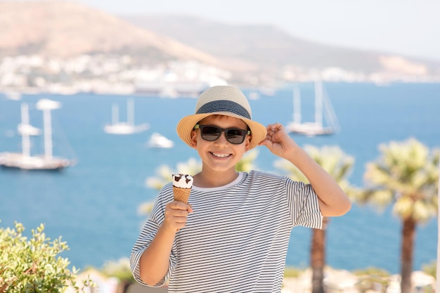 Portrait of happy kid with sea on background Handsome smiling boy in straw hat and sunglasses eating ice cream outdoor having fun at summer vacation