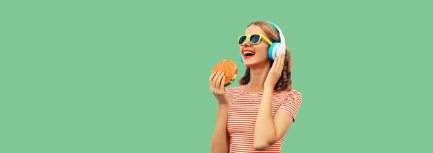 Photo portrait of happy laughing young woman listening to music in headphones holding burger fast food