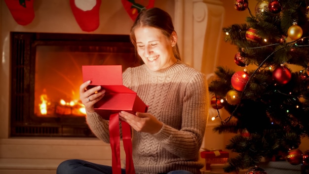 Portrait of happy laughing young woman looking inside of glowing Christmas gift box while sitting on floor next to the fireplace