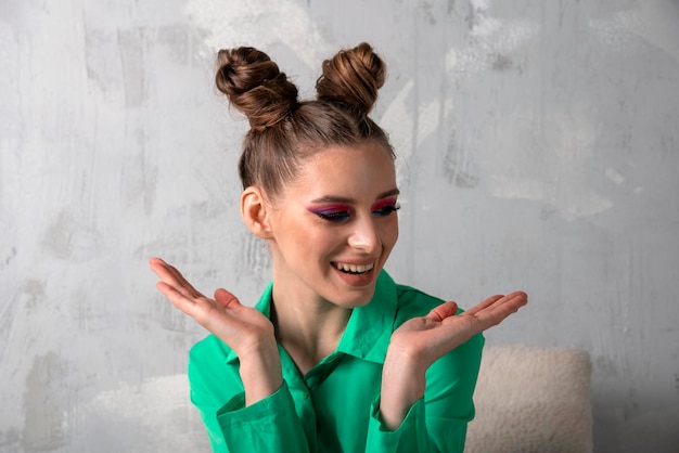 Portrait of happy laughing young woman with bright makeup and bun hairstyle Green shirt Gray background