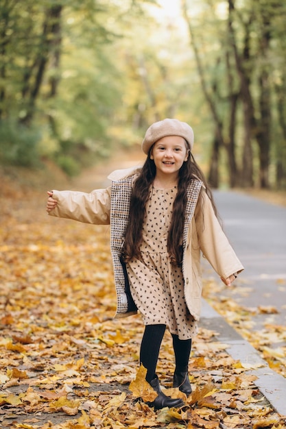 Portrait of happy little girl in beige coat and beret holding yellow maple leaves and spending time in autumn park