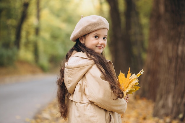 Portrait of happy little girl in beige coat and beret holding yellow maple leaves and spending time in autumn park