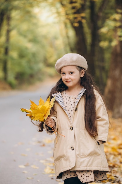 Portrait of happy little girl in beige coat and beret holding yellow maple leaves and spending time in autumn park