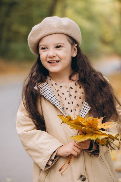 Portrait of happy little girl in beige coat and beret holding yellow maple leaves and spending time in autumn park