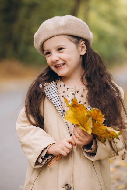 Portrait of happy little girl in beige coat and beret holding yellow maple leaves and spending time in autumn park