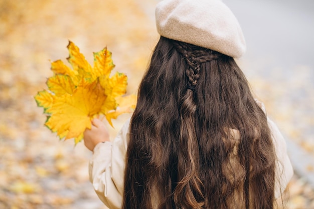 Portrait of happy little girl in beige coat and beret holding yellow maple leaves and spending time in autumn park