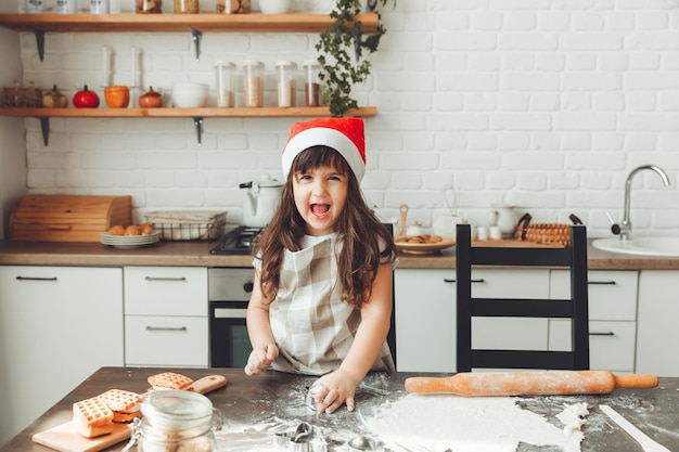 Portrait of a happy little girl in a Santa hat rolling out dough on the kitchen table a child preparing Christmas cookies