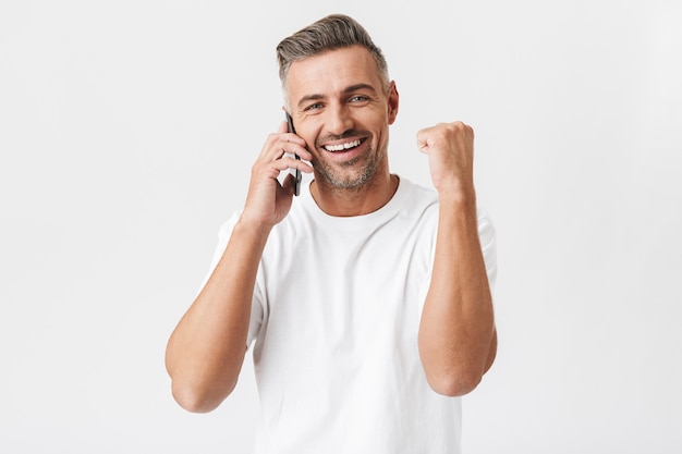 Portrait of happy man 30s wearing casual t-shirt holding smartphone and having mobile conversation isolated on white