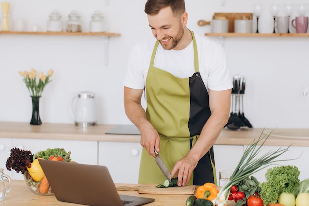 Portrait of happy man at home man cooking vegetable salad looking at camera and smiling slicing vegetables using laptop for online cooking training