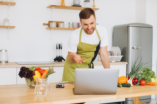 Portrait of happy man at home man cooking vegetable salad looking at camera and smiling slicing vegetables using laptop for online cooking training