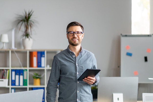 Portrait of happy mature office worker holding digital tablet looking and smiling at camera standing