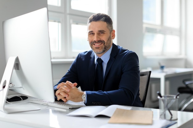 Portrait of happy middle aged businessman sitting in front of computer at workplace in office