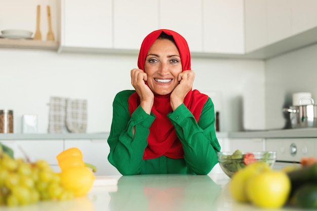 Portrait of happy middle aged muslim housewife posing in kitchen sitting at table and smiling at camera free space