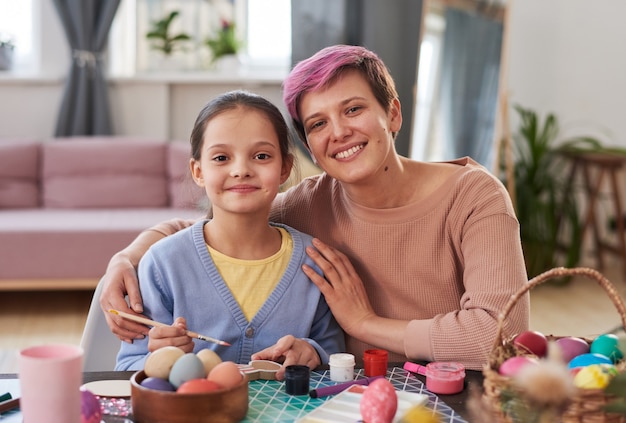 Portrait of happy mother embracing her daughter and smiling at camera while painting Easter eggs at the table