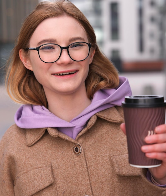 Portrait of happy positive beautiful teen teenager girl young woman student in glasses is presenting holding cup of tea beverage or coffee to go in hand walking outdoors in coat Vertical photo