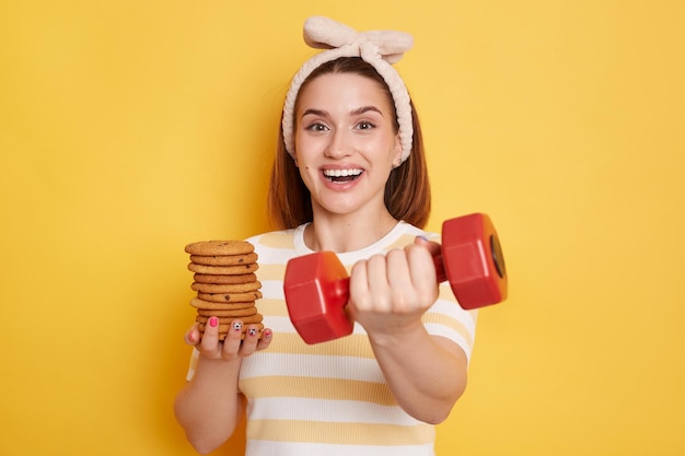 Portrait of happy positive woman wearing striped shirt and hair band holding out red dumbbell and cookies offering workout and eat whatever you want posing isolated over yellow background