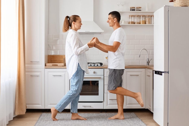 Portrait of happy positive young adult Caucasian man and woman, posing indoor, dancing in kitchen, celebrating their anniversary, wearing casual style attires.