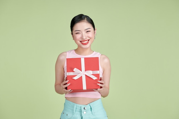 Portrait of a happy pretty asian woman holding present box while standing isolated over green pastel background