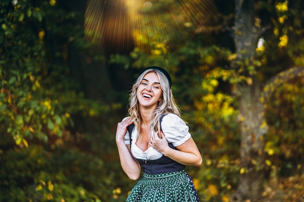 Portrait of happy pretty blond girl in dirndl, traditional beer festival dress, standing outdoors with blured colorful trees behind