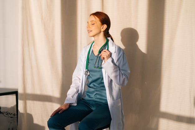 Portrait of happy pretty female doctor in white coat sitting on chair near window in sunny day in medical clinic office.