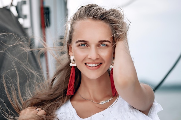 Portrait of happy pretty girl with white dress, long red earrings and long curly blonde hair standing on yacht at summertime. looking at camera with toothy smile. close UP