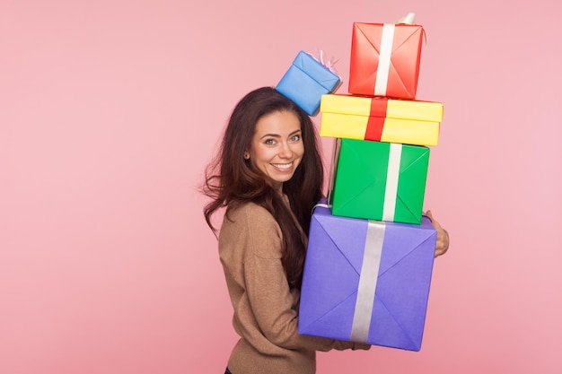 Portrait of happy pretty young woman smiling pleased to camera and holding mount of gift boxes enjoying many best presents celebrating birthday indoor studio shot isolated on pink background