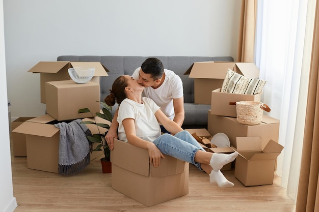 Portrait of happy romantic couple having fun while moving into new house attractive woman sitting in cardboard box while man kissing her with love na gentle