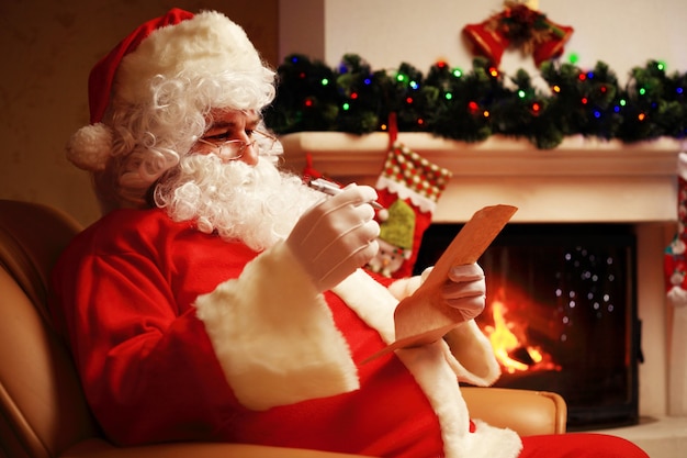 Portrait of happy Santa Claus sitting at his room at home near Christmas tree and big sack and reading Christmas letter or wish list