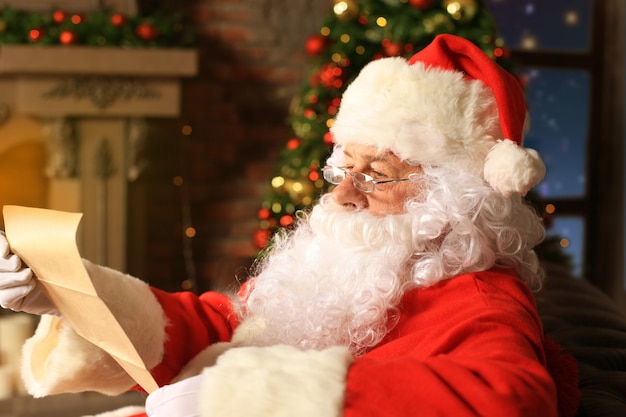 Portrait of happy Santa Claus sitting at his room at home near Christmas tree and reading Christmas letter or wish list.