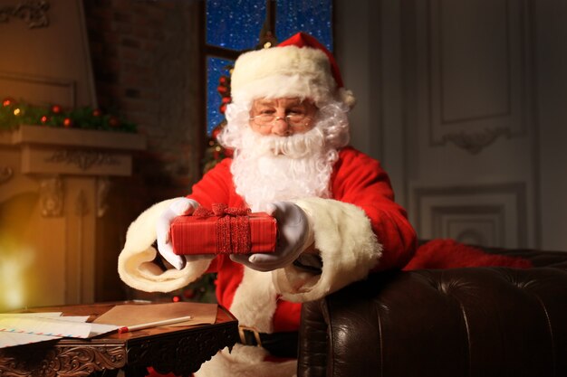 Portrait of happy Santa Claus sitting at his room at home near Christmas tree and reading Christmas letter or wish list.