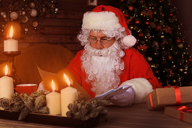 Portrait of happy Santa Claus sitting at his room at home near Christmas tree and reading Christmas letter or wish list.